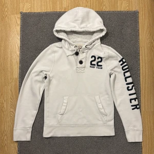 Vit Vintage Hollister Hoodie Med Knappar - Sällsynt vintage Vit hoodie från Hollister med broderat '22 Surf Team' på bröstet och stort Hollister-tryck längs ena ärmen. Tröjan har huva med snörning, knappar vid halsen och en klassisk känguruficka. (Små svarta prickar finns på hoodien men syns knappast då vintage hoodies har sliten/använd design, annars är det bara att tvätta hoodien!). Perfekt för dig med avslappnad stil! 