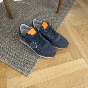 Blå Philippe Model sneakers med orange detaljer - Snygga sneakers från Philippe Model i mörkblå mocka och mesh med coola orange detaljer på plös och sula. Skorna har snörning och en vit platt sula, perfekt för en sportig och trendig look.