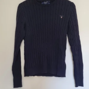 Mörkblå kabelstickad tröja från GANT - Kabelstickad mörkblå tröja från GANT i stretchig bomull. Klassisk rund halsringning och diskret logga broderad på bröstet. Snygg ribbad kant vid ärmslut och nederkant. Perfekt för lager-på-lager och stilren vardagslook. 