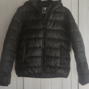 Svart pufferjacka från Champion XL (barn) - Svart pufferjacka från Champion i storlek XL med huva och dragkedja. Jackan har en quiltad design och är perfekt för kyliga dagar. Snygg och enkel stil som funkar till det mesta. Klassisk logga på bröstet och två fickor framtill.