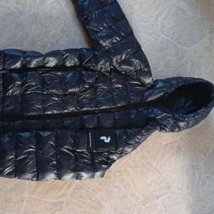 Svart dunjacka Peak Performance - Säljer en svart, glansig dunjacka från Peak Performance med huva och klassisk quiltad design. Jackan har en stor logotyp på ärmen och dragkedja framtill. Perfekt för kalla dagar när du vill hålla stilen och värmen. Två små hål som borde gå att sy.