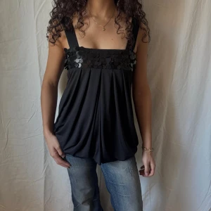 Svart festtopp med paljettdetalj - Svart topp med breda axelband och lös passform. Toppen har en snygg paljettrad över bysten som ger extra glans. Tillverkad i ett mjukt, lätt material som faller snyggt. Perfekt för dig som vill sticka ut lite extra.