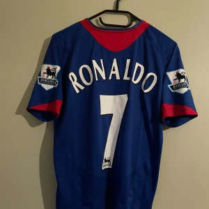 Manchester United Ronaldo 7 bortatröja - Blå Manchester United bortatröja med röda detaljer, Ronaldo 7-tryck på ryggen och Barclays Premier League-patchar på ärmarna. Klassisk klubbmärke och Vodafone-logga framtill. Perfekt för dig som älskar fotboll och retrostil.