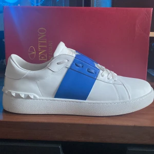 Valentino Garavani Open sneakers vit/blå - Säljer ett par Valentino Garavani Open sneakers i vitt skinn med bred blå rem över mitten. Klassisk chunky sula med nitar baktill och rund tå. Snygg och stilren design med vita snören och diskret logga på plösen. Perfekta för dig som vill sticka ut med lyxig streetstyle.