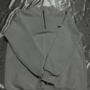Grå half zip sweatshirt från Lacoste - Säljer en stilren grå sweatshirt från Lacoste med half zip och långärmad design. Tröjan har en liten krokodillogga på bröstet och ribbade muddar vid ärmslut och nederkant. Perfekt för dig som gillar enkel och clean stil.