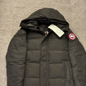 Canada Goose jacka - Säljer en svart dunjacka från Canada Goose med huva och klassisk logga på ärmen. Jackan har quiltad design, dragkedja framtill och är perfekt för kalla dagar. Snygg och stilren modell som håller dig varm hela vintern. (Helt ny)