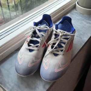 Adidas vita och blå fotbollsskor - Säljer ett par Adidas fotbollsskor i vitt med blå och röda detaljer. Skorna har snörning, rund tå och platt sula med dobbar för gräsplan. Ovandelen är i syntetmaterial och insidan är fodrad i blått tyg. Perfekta för dig som vill sticka ut på planen! De är lite slitna på framsidan