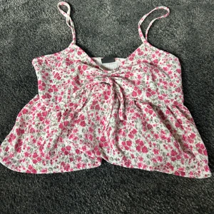 Blommig linnetopp från Gina Tricot - Supersöt linnetopp från Gina Tricot i storlek M. Toppen är vit med rosa och gröna blommor, har tunna axelband och en snygg knyt-detalj framtill. Materialet är lätt och luftigt, perfekt för varma dagar. Passformen är loose och toppen har en volang nedtill.