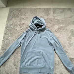 Ljusblå hoodie från Polo Ralph Lauren - Säljer en stilren ljusblå hoodie från Polo Ralph Lauren med klassisk huva och snörning. Tröjan har en broderad logga på bröstet, känguruficka och är tillverkad i mjuk bomull. Perfekt för chill dagar och enkel att matcha med jeans eller joggers.