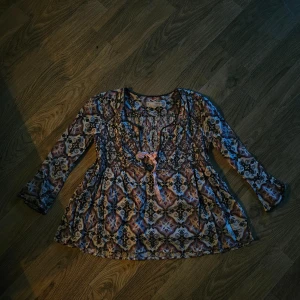 Mönstrad blus från Odd Molly med rosett - Säljer en bohemisk blus från Odd Molly med färgstarkt mönster i rosa, blått, beige och svart. Blusen har långärmade, lätt utsvängda ärmar och en söt ljusrosa rosett framtill. Osäker på storlek, S eller M förmodligen 