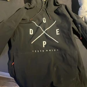 Dope skidjacka  - Svart vindjacka från Dope med stor vit logga på bröstet, huva med snörning och dragkedja på ärmen. Jackan har rödsvarta detaljer och är fodrad med mjuk fleece på insidan. Perfekt för dig som gillar streetwear och vill ha en snygg och funktionell jacka.