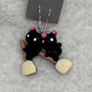 Halloween örhängen katter  - Unika örhängen med två svarta katter med rosa öron som sitter på en brun kvast med beige borste. Perfekt för dig som gillar gulliga och annorlunda smycken med djurmotiv. Hängande modell med silvriga krokar.