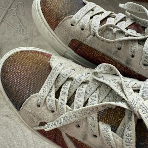 Glittriga sneakers från Zadig & Voltaire - Säljer ett par unika sneakers från Zadig & Voltaire med glittrig ovandel i brons, guld och röd ton. Skorna har ljusgrå mockadetaljer, vit sula och cool blixtdetalj på sidan. Skosnörena är grå med textprint. Perfekta för dig som vill sticka ut.