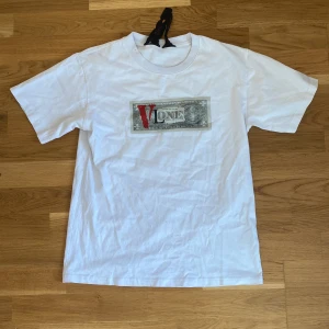 Vit VLONE t-shirt med dollartryck - Vit t-shirt från VLONE i 100% bomull med unikt dollartryck och röd V-logga på bröstet. Stor V-grafik med ansikte på ryggen. Klassisk passform och crewneck. Grym streetwear-vibe och tydlig branding, perfekt för dig som gillar statement-plagg.