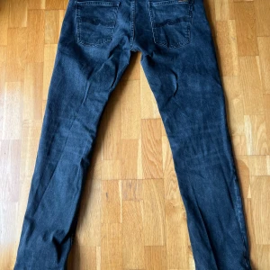 Grå/svarta Nudie jeans W32 L34 - Snygga grå/svarta jeans från Nudie Jeans med klassisk femficksdesign och raka ben. Jeansen har normal passform och är tillverkade i mjukt denimtyg som ger en skön känsla. Perfekta för dig som gillar en enkel och stilren look. Knappen är utbytt men kan bytas gratis hos Nudie. Model Grim Tim 