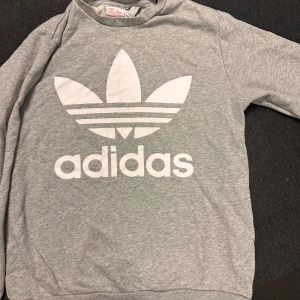 Grå sweatshirt från Adidas - Säljer en grå sweatshirt från Adidas med den klassiska vita trefoil-loggan tryckt framtill. Tröjan har rund halsringning och långa ärmar, perfekt för en sportig och avslappnad stil.