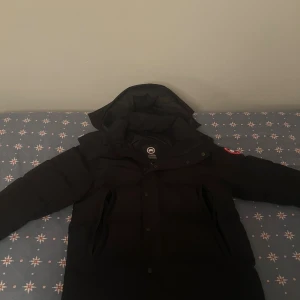 Svart dunjacka från Canada Goose - Säljer en svart dunjacka från Canada Goose med klassisk patch på ärmen. Jackan har huva, dragkedja och tryckknappar framtill. Fodrad insida och puffig design som håller dig varm under kalla dagar. Perfekt för dig som vill ha en stilren och funktionell vinterjacka. Päls och påse kommer med jackan. 