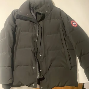 Svart dunjacka från Canada Goose - Säljer en svart dunjacka från Canada Goose med klassisk patch på ärmen och avtagbar huva med päls. Jackan har dragkedja, tryckknappar och flera fickor. Perfekt för kalla vinterdagar och riktigt snygg streetstil. Vill informera om en liten riva i insidan av jackan vid höger arm, men det är såklart inget som påverkar hur den används