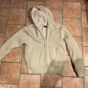 Fodrad cardigan från Schott NYC - Säljer en vit stickad hoodie från Schott NYC med mjukt foder på insidan. Tröjan har ribbade detaljer på ärmar och nederkant, dragkedja framtill och huva med snörning. Perfekt för kyliga dagar och har en clean, stilren look.