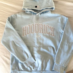 Hoodrich hoodie, strl M - Säljer en ljusblå hoodie från Hoodrich med stor vit logga på bröstet, tryck på ryggen och huvan. Tröjan har huva, magficka och ribbade muddar. Materialet är mjuk bomull och passformen är normal. Tröjan är använd men i väldigt fint skick. Tröjan är i storlek M, passar både dam och herr. 