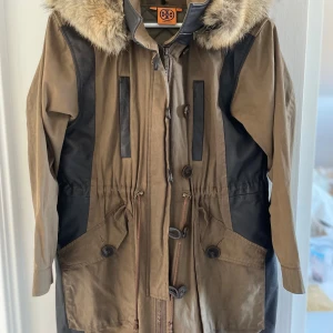 Parkajacka med päls från Tory Burch - Snygg parkajacka från Tory Burch i brunt och svart med päls på huvan. Jackan har stora fickor, knappar, dragkedja och snörning i midjan. Materialmix av bomull och detaljer i skinnimitation ger en cool kontrast. Perfekt för kyliga dagar.