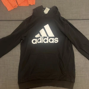 Svart Adidas hoodie med stor logga - Svart hoodie från Adidas i storlek  med stor vit Adidas-logga på bröstet. Klassisk huva och magficka framtill, tillverkad i mjuk bomull och polyesterblandning. Perfekt för dig som gillar sportig och chill stil.