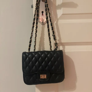 Svart quiltad axelväska med kedja - Snygg svart axelväska i quiltad design med guldfärgade detaljer och kedjerem. Väskan har ett klassiskt lock med vridlås och ett innerfack med dragkedja. Perfekt storlek för det viktigaste och ger en lyxig känsla till din outfit.