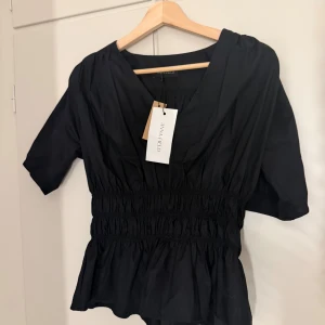 Svart blus med v-ringning från Anna Field - Svart blus från Anna Field med v-ringning och korta ärmar. Toppen har smockad midja som ger en snygg siluett och lätt volang nertill. Tillverkad i ett mjukt bomullstyg, perfekt för dig som gillar stilrena och bekväma plagg.