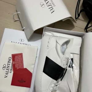 Valentino Garavani Open sneakers vit/svart - Snygga Valentino Garavani Open sneakers i vitt skinn med bred svart läderdetalj över mitten. Klassisk rund tå, platt sula och ikoniska nitar baktill. Perfekta för dig som vill ha en clean och lyxig look med streetkänsla.KVITTO FINNS