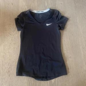 Svart Nike Pro t-shirt med v-ringning - Nike Pro t-shirt i svart med vit logga på bröstet. T-shirten har v-ringad hals och korta ärmar, tillverkad i ett stretchigt och lätt polyestermaterial som andas. Perfekt för dig som gillar sportig och clean stil.