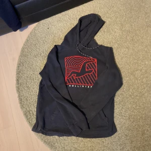 Marinblå hoodie från Hollister med röd logga - Marinblå hoodie från Hollister med stor röd grafisk logga och fågel på bröstet. Tröjan har huva med snörning och är långärmad. Perfekt för dig som gillar en stilren men ändå cool look.