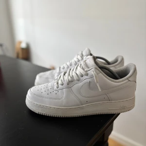 Nike Air Force 1 vita sneakers - Fräscha vita Nike Air Force 1 sneakers med klassisk låg siluett och rund tå. Skorna har snörning, perforerade detaljer på tån och är tillverkade i skinn med en platt sula. Perfekt för dig som gillar stilrena och tidlösa sneakers.