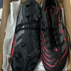 Adidas Predator Club fotbollsskor 43 1/3 - Adidas Predator Club FG/MG fotbollsskor i svart med röda och grå detaljer. Snyggt mönstrad ovandel med snörning och platt sula med dobbar för bra grepp på planen. Skorna har en sportig och modern look, perfekta för dig som vill sticka ut på fotbollsplanen.
