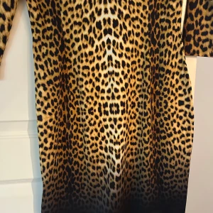 Leopardmönstrad klänning Just Cavalli - Snygg leopardmönstrad klänning från Just Cavalli med coola smyckesdetaljer upptill. Klänningen har trekvartsärm, rund halsringning och är figurnära. Materialet är stretchigt och bekvämt, perfekt för dig som vill sticka ut med ett djurmönster och unika detaljer.