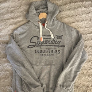 Grå hoodie från Superdry med tryck - Säljer en grå hoodie från Superdry med svart tryck framtill och vita snören i huvan. Klassisk känguruficka och ribbade muddar. Perfekt för dig som gillar en avslappnad och stilren look.