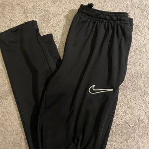Svarta Nike Dri-FIT träningsbyxor  - Svarta träningsbyxor från Nike med Dri-FIT-teknologi och klassisk vit swoosh-logga på benet. Byxorna har resår i midjan och är tillverkade i ett lätt och funktionellt syntetmaterial som andas. Perfekta för träning eller chill. (Passar även S, då de är lite mindre i storleken!)
