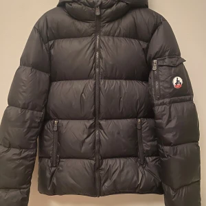 Svart pufferjacka från JOTT - Svart pufferjacka från JOTT med huva och flera dragkedjeförsedda fickor. Jackan har ett snyggt JOTT-märke på ärmen och quiltad design. Perfekt för kalla dagar, med en modern och clean look. Insidan har coolt tryck med illustrationer. Nypris 3799 kr!