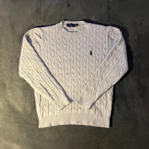 Ljusgrå kabelstickad tröja Polo Ralph Lauren - Kabelstickad ljusgrå tröja från Polo Ralph Lauren med klassisk rund hals och broderad logga på bröstet. Tröjan är i bomull och den är i bra skick och det finns inga synliga defekter. Tröjan är i storlek M.
