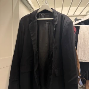 Svart kavaj från Pull&Bear - Stilren svart oversize kavaj från Pull&Bear med klassisk krage och två fickor framtill. Enkelknäppt modell med slät yta och diskret passform. Perfekt för dig som vill ha en clean och tidlös look.