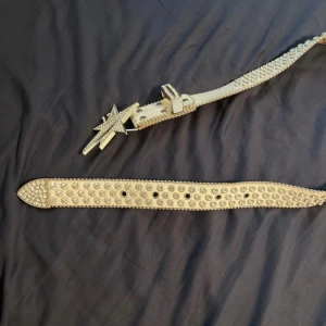 Lukamachain bälte - Säljer ett vitt bälte med glittriga stenar och ett stort stjärnformat spänne med siffran 12. Bältet har krokodilpräglad yta och är dekorerat med silverfärgade nitar och kristaller längs hela längden. Perfekt för dig som gillar att sticka ut med accessoarer.