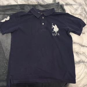 Mörkblå pikétröja från U.S. Polo Assn. - Mörkblå pikétröja med klassisk krage och knappar framtill från U.S. Polo Assn. Stor vit broderad logga på bröstet och en stor vit siffra 3 på ärmen. Skön och stilren, perfekt till jeans eller shorts.