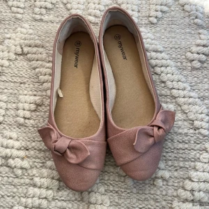 Rosa ballerinaskor med rosett från MyWear - Säljer ett par söta ballerinaskor i ljusrosa från MyWear, storlek 38. Skorna har rund tå, platt sula och en fin rosettdetalj framtill. Materialet är syntet med lätt textur och insidan är mjuk för extra komfort. Perfekt för dig som gillar en feminin och enkel stil.