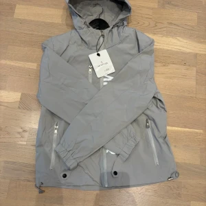 Grå vindjacka från Moncler med huva - Snygg grå vindjacka från Moncler med huva och stora vita logotypdetaljer framtill. Jackan har tre dragkedjeförsedda fickor, justerbar nederkant och Moncler-märke på ärmen. Perfekt för dig som vill ha en stilren och sportig look. Tveka inte på att DM'a för frågor. Pris ej hugget i sten ( FINNS I SVART OCKSÅ )