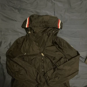 Svart vindjacka från M - Svart vindjacka från M med huva och dragsko, dekorerad med röda och vita ränder på huvan. Jackan har dragkedja framtill och klassisk Moncler-logga på ärmen. Perfekt för dig som vill ha en stilren och sportig look. Passar både S och M