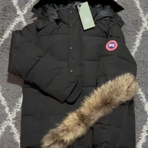 Canada Goose jacka - Svart dunjacka från Canada Goose med klassisk patch på ärmen, avtagbar huva och snygg fuskpälsdetalj. Jackan är quiltad och har knappar framtill. Perfekt för kalla vinterdagar och riktigt stilren look.