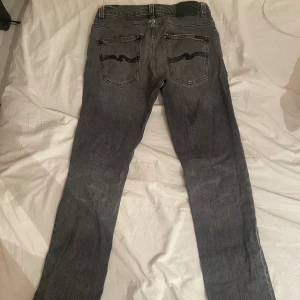 Grå jeans från Nudie Jeans grim Tim  - Snygga grå jeans från Nudie Jeans med klassisk svart broderad våg på bakfickorna. Jeansen har fem fickor, raka ben och normal passform. Tillverkade i mjukt denimtyg som ger en avslappnad vibe. Perfekta för dig som gillar stilrena och tidlösa jeans.