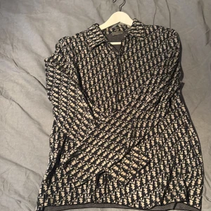 Dior skjorta  - Säljer en snygg långärmad skjorta från Dior med ikoniskt mönster i mörkblått och beige. Skjortan har klassisk krage och knappar framtill. Materialet känns lätt och lyxigt, perfekt för dig som gillar statement-plagg med designer-vibe.