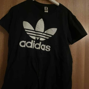 Svart Adidas t-shirt med stor logga - Svart t-shirt från Adidas med den klassiska stora treklöver-loggan i vitt framtill. Modellen är kortärmad och har rund hals. Materialet är mjuk bomull som är skön mot huden. Perfekt för dig som gillar sportiga och stilrena plagg med tydlig branding.