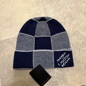 Rutig mössa från Louis Vuitton - Snygg rutig mössa från Louis Vuitton i marinblått och grått. Mössan har ett coolt rutmönster och broderad text framtill. Tillverkad i lyxig mix av kashmir, ull och silke för extra mjuk känsla. Perfekt för dig som vill ha en exklusiv och trendig look.
