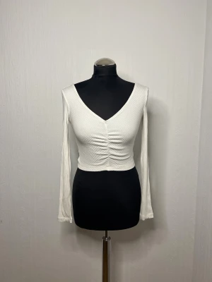 Vit ribbad croppad topp XS - Säljer en vit croppad topp i ribbad bomull med lång ärm och snygg v-ringning. Toppen har rynkade detaljer framtill som ger en cool look. Perfekt för dig som gillar stilrena och trendiga plagg med lite edge. V ringad även bak på ryggen. 